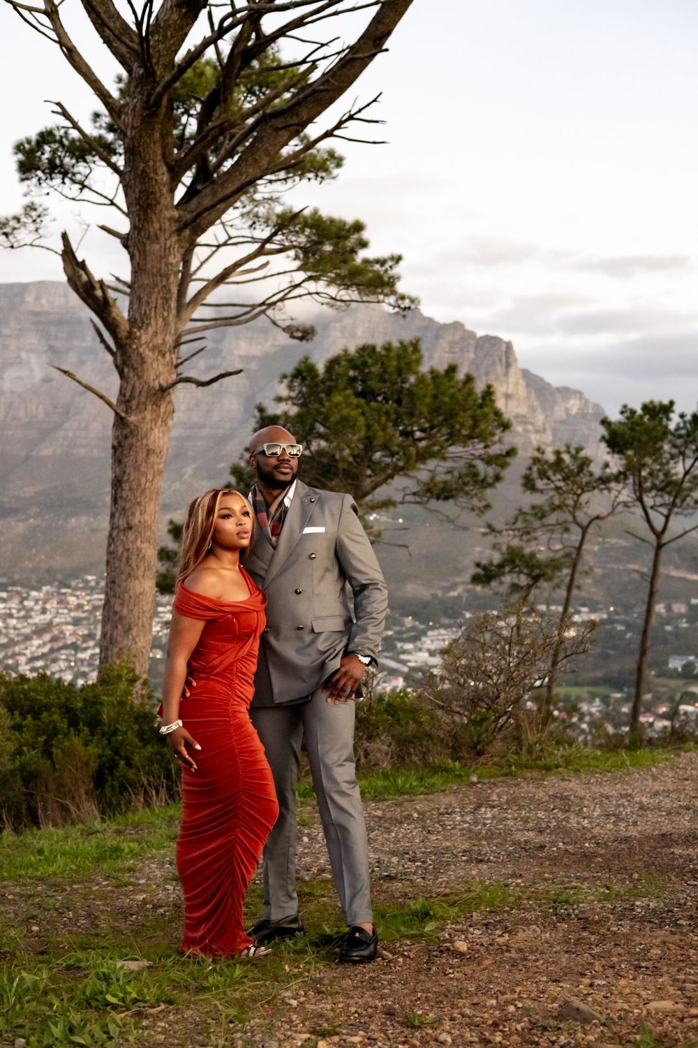 A couple in formal attire poses on a mountain overlook with Table Mountain and Cape Town in the background.