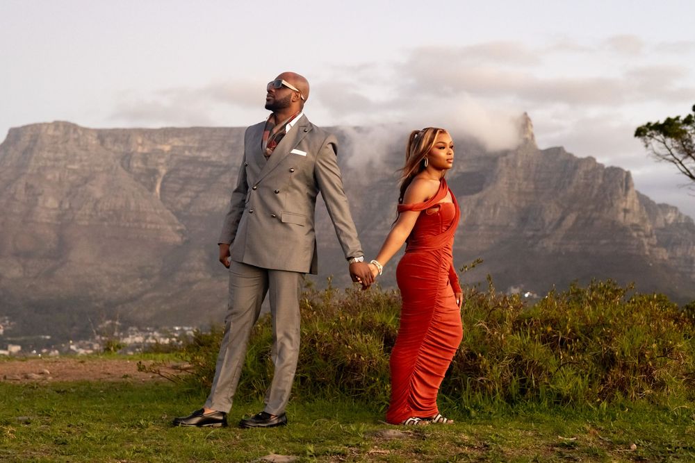Couple in formal attire holds hands while posing against dramatic mountain backdrop in Cape Town, South Africa.