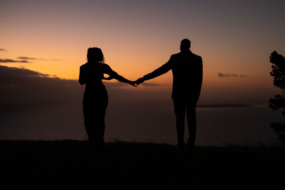 Silhouetted couple holds hands against dramatic orange sunset sky over horizon.