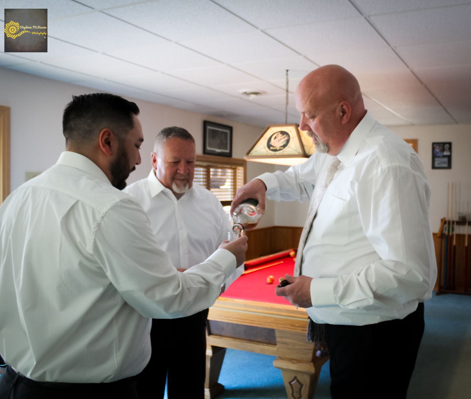 Three people in white dress shirts gathered around a pool table in a room with wood paneling and overhead lighting.
