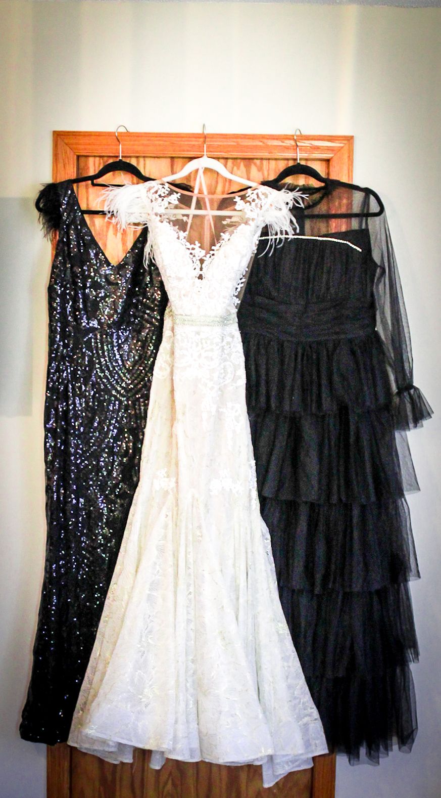 Three formal dresses hanging on a wooden door - a black sequined gown, white lace wedding dress, and black tiered tulle dress.