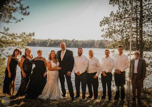 Wedding party in formal attire poses together on scenic lakeside cliff at sunset with pine trees in background.