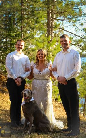 Wedding group in white attire poses outdoors with a brown dog against a backdrop of tall pine trees and a lake.