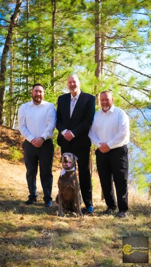 A chocolate Labrador dog sits on a wooded hillside with three people in formal attire among tall pine trees.