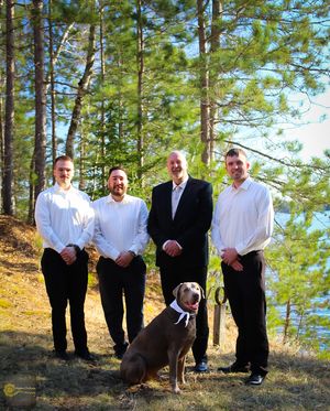 Four men and a large dog pose outdoors on a grassy hillside with pine trees in the background.