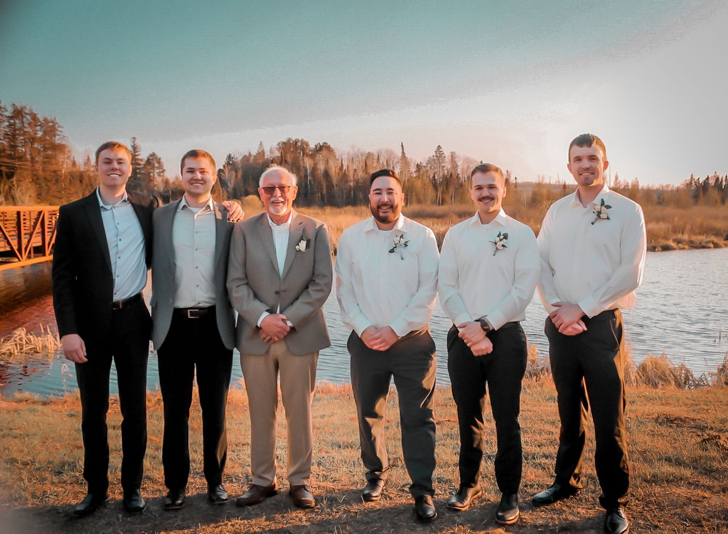 A wedding party poses together in front of a scenic lakeside setting during sunset with a warm autumn glow.