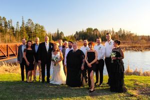 Wedding party stands outdoors in formal attire with a scenic lake and forest backdrop during golden hour sunset.