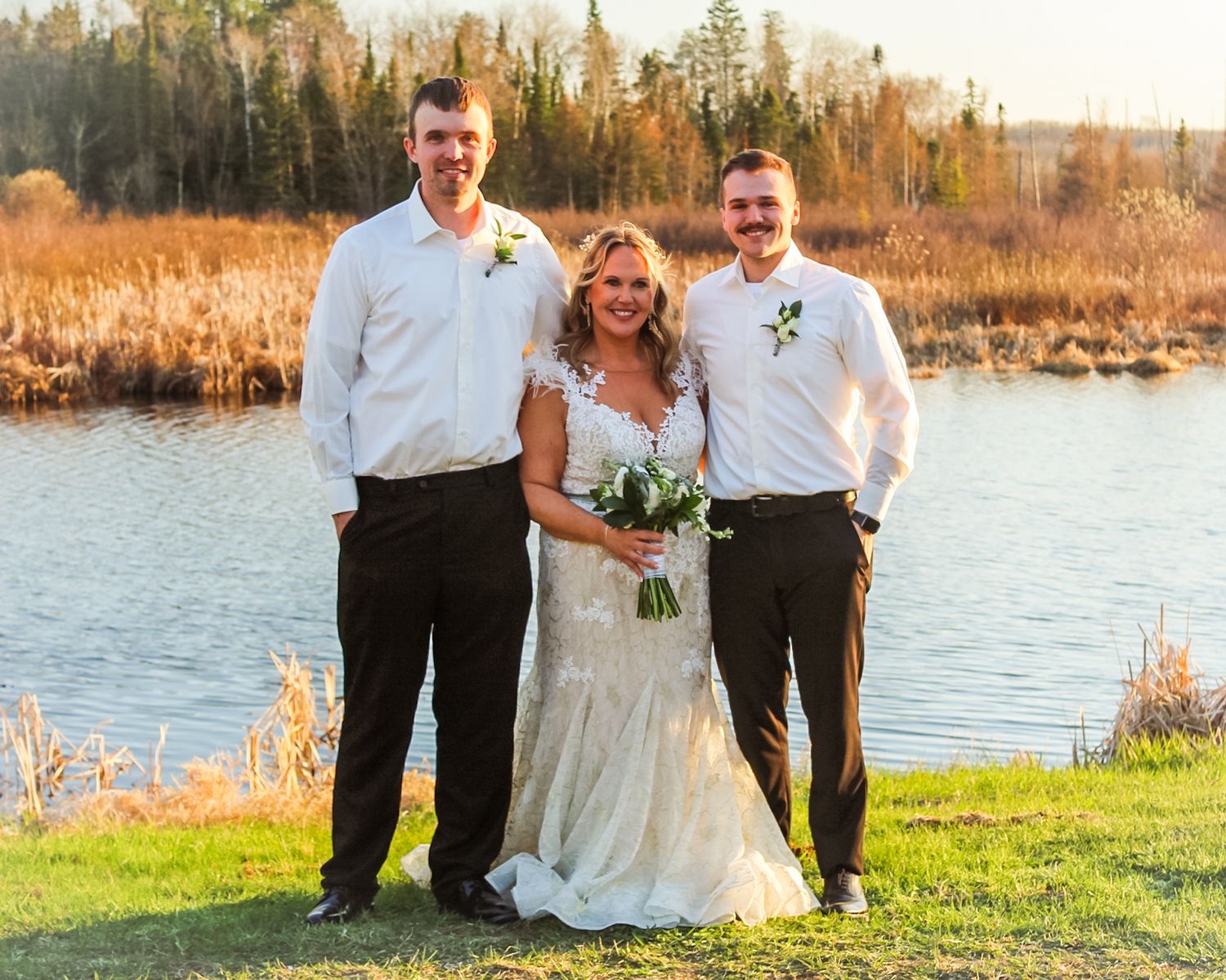 Three people pose together on grassy shore by a lake at sunset with fall foliage and trees in background.