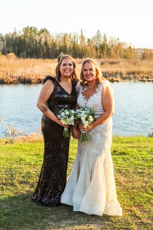 Two people in formal gowns stand beside a lake at sunset holding white flower bouquets on green grass.