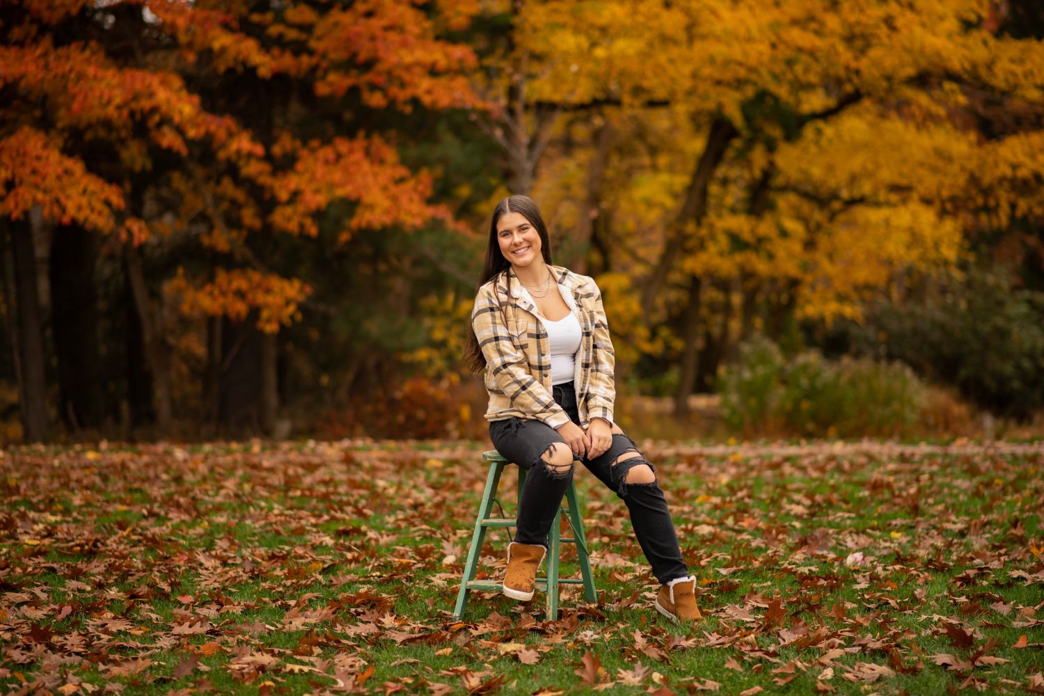 Someone sits on a stool in a plaid shirt surrounded by colorful autumn foliage and fallen leaves in a forest setting.