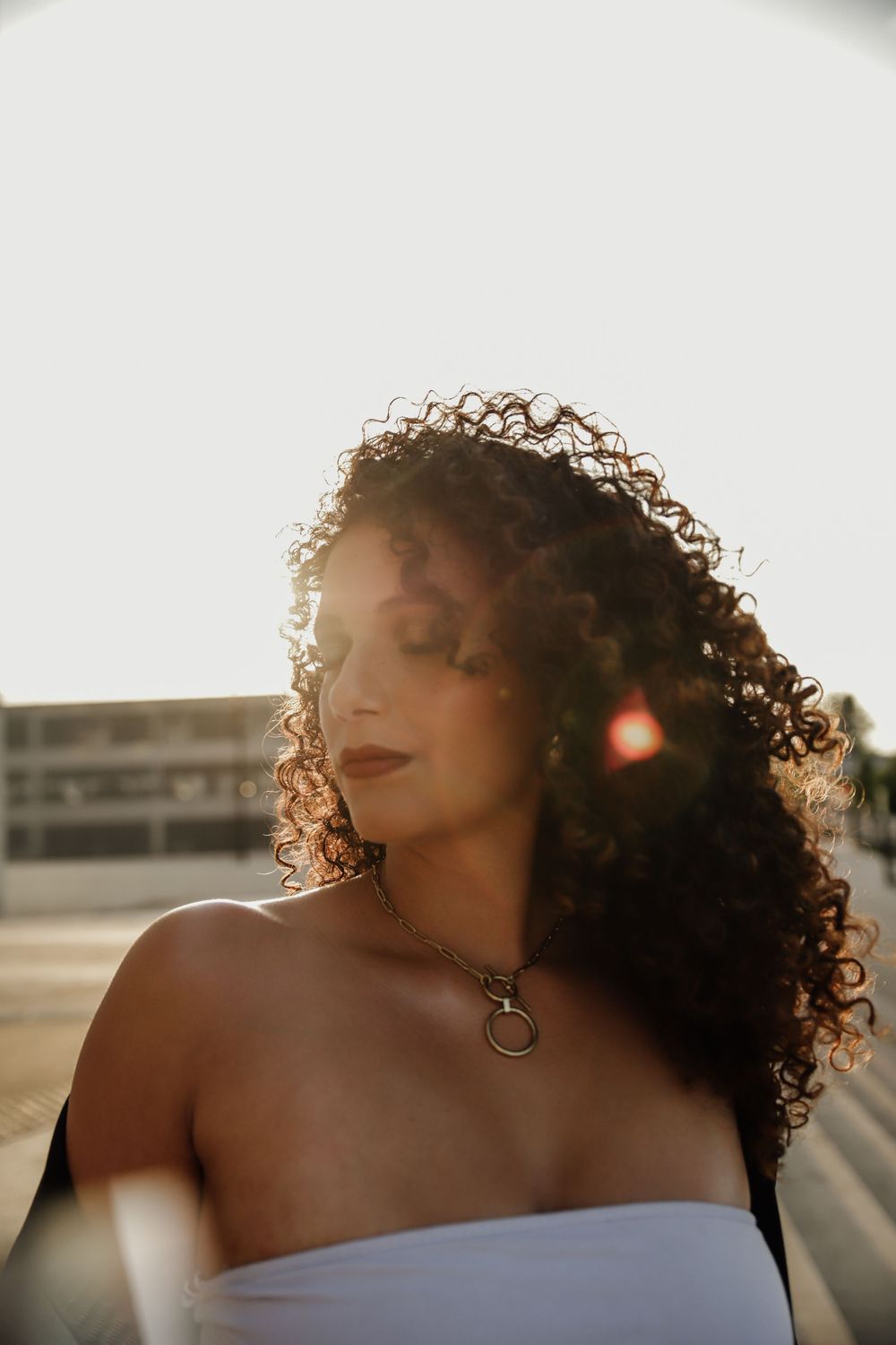 A person with curly hair in a white top poses in golden hour sunlight, with a circular necklace pendant.