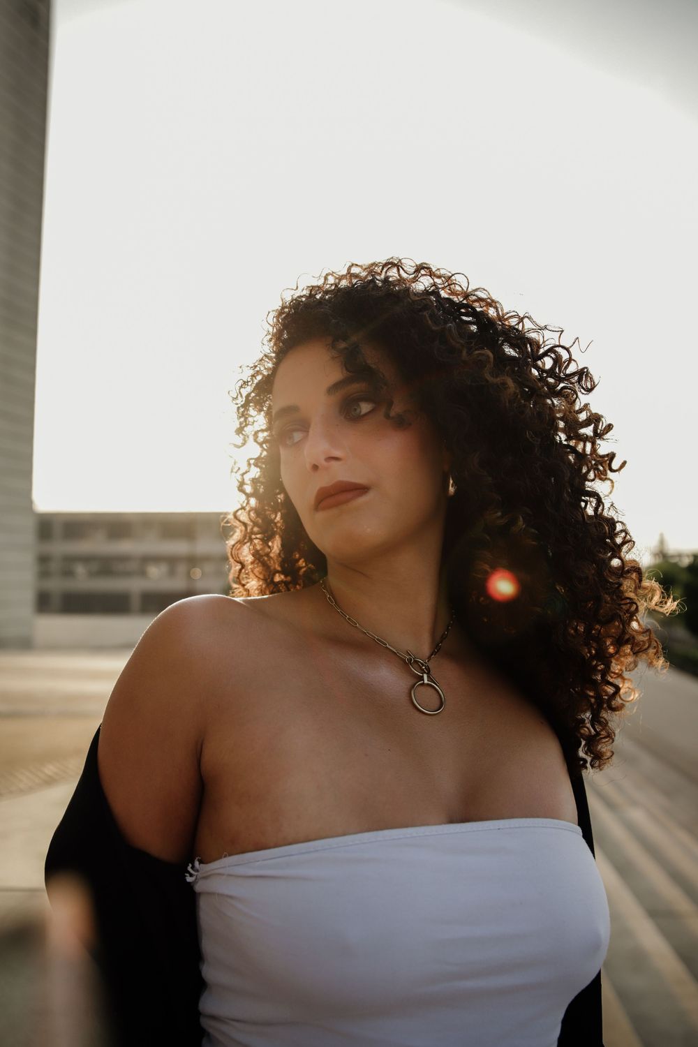 Portrait in golden sunlight capturing curly hair, white off-shoulder top and pendant necklace with warm sunset lighting.