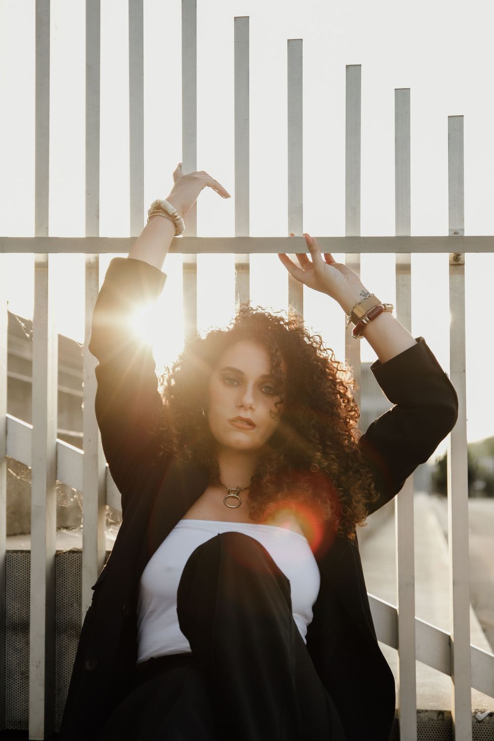 A dramatic fashion portrait with glowing sunlight through metal bars, featuring black jacket and white top against sunset.