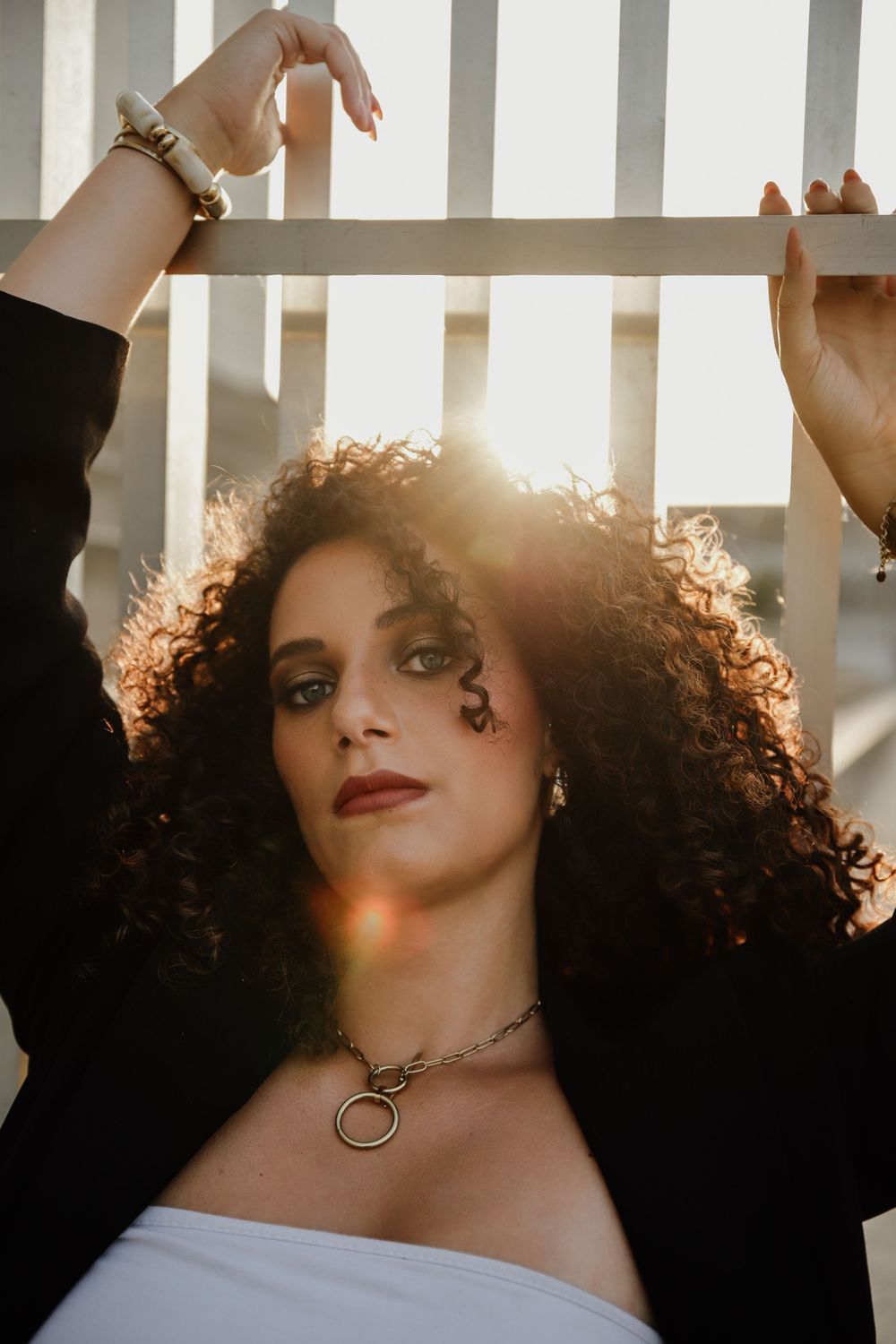 Sun-drenched portrait featuring curly hair, red lipstick, and a circular pendant necklace against white railings.