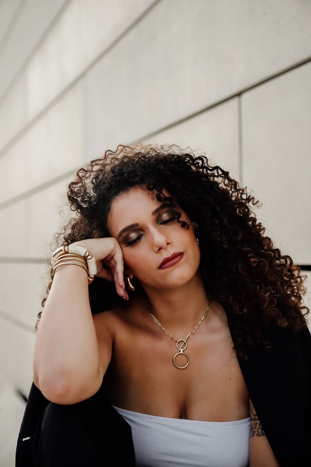 Portrait against white wall showing curly dark hair, white top, and layered necklaces in warm lighting.
