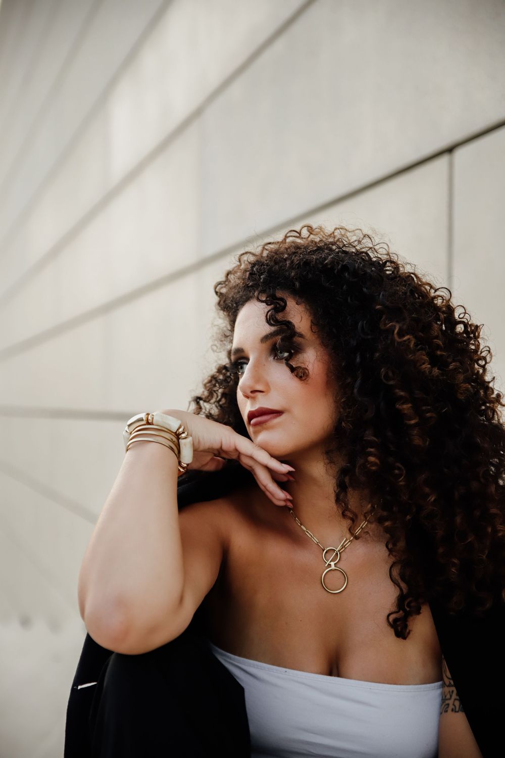 A person with curly hair and flowing dress poses against a white wall in an artistic photoshoot series.