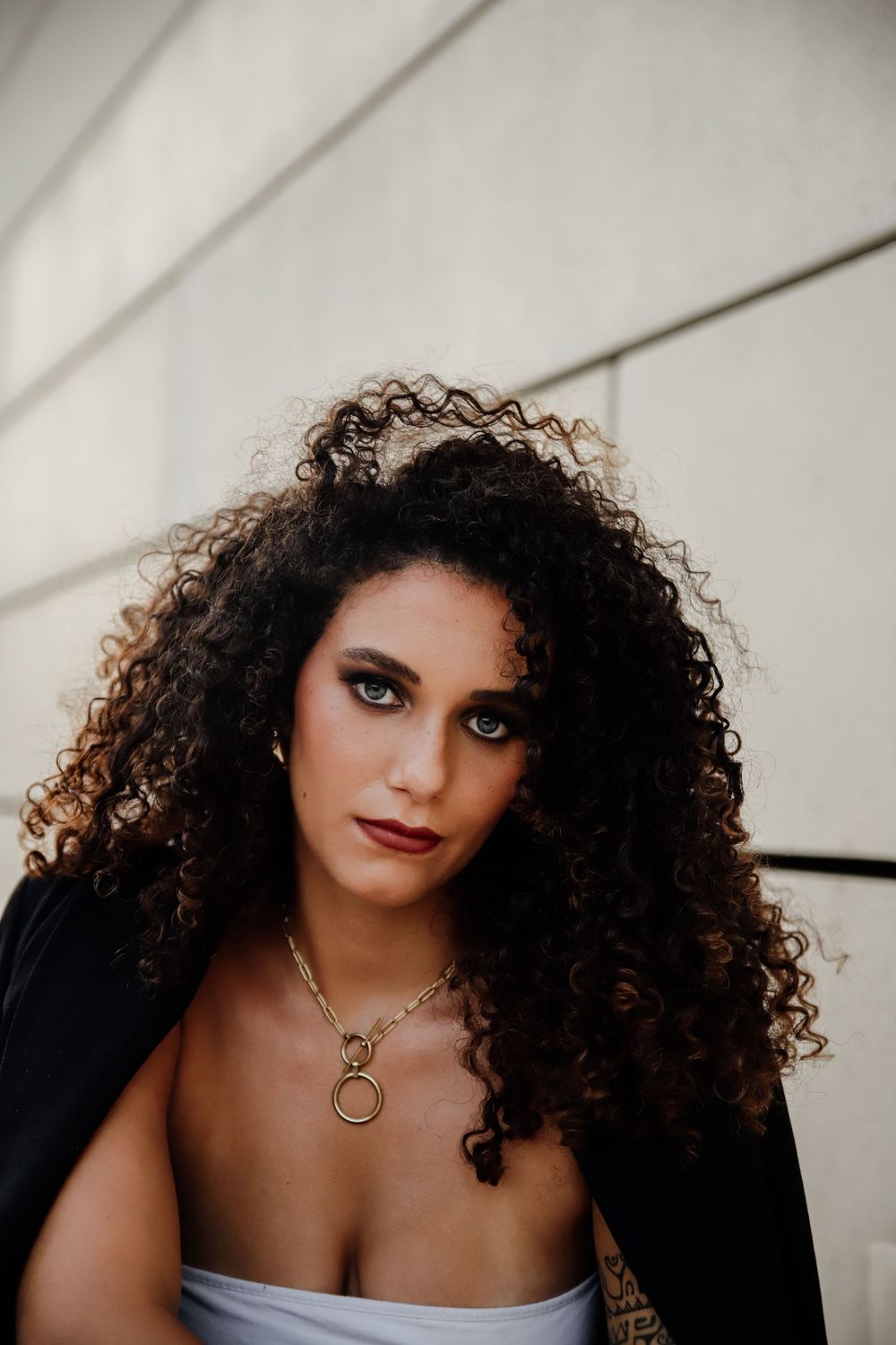 Person with curly dark hair and dramatic eye makeup poses against a white textured wall in moody lighting portraits.