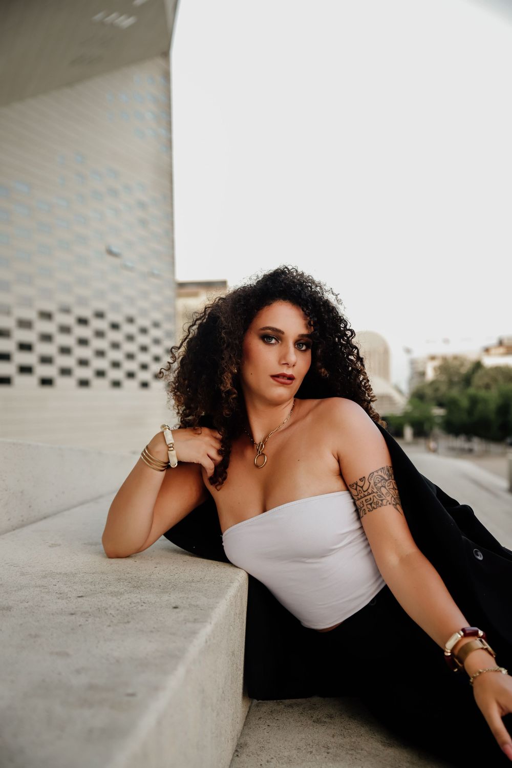 A woman in a white dress and black jacket poses dramatically against a textured concrete wall.