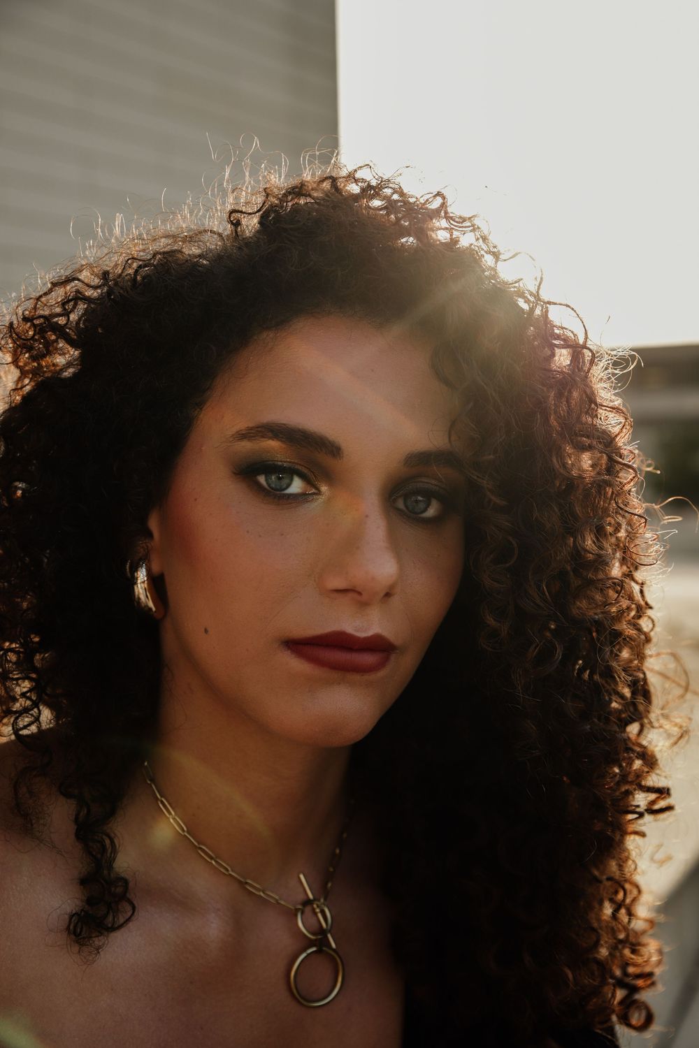 A portrait photo illuminated by golden sunlight highlighting natural curly hair, red lipstick and a gold necklace.