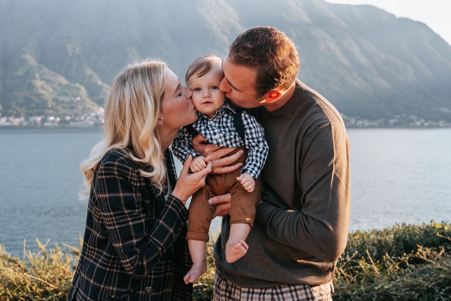 Giovane coppia di genitori bacia le guance del loro bambino, la mamma da un lato e il papà dall'altro. Sullo sfondo le rive del Lago di Como