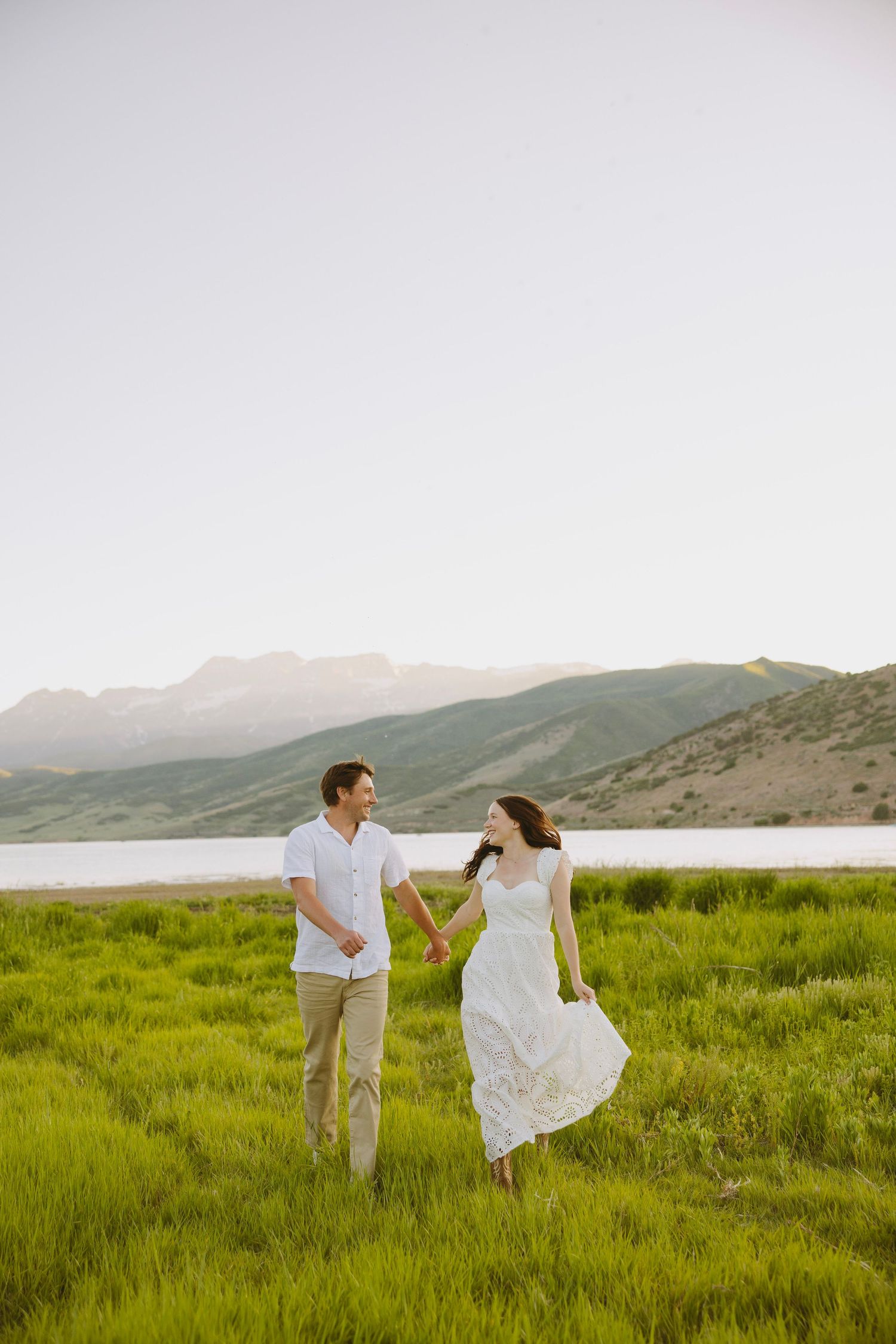 A bride in a flowing white dress stands gracefully on a wooden deck surrounded by evergreen mountains at sunset.