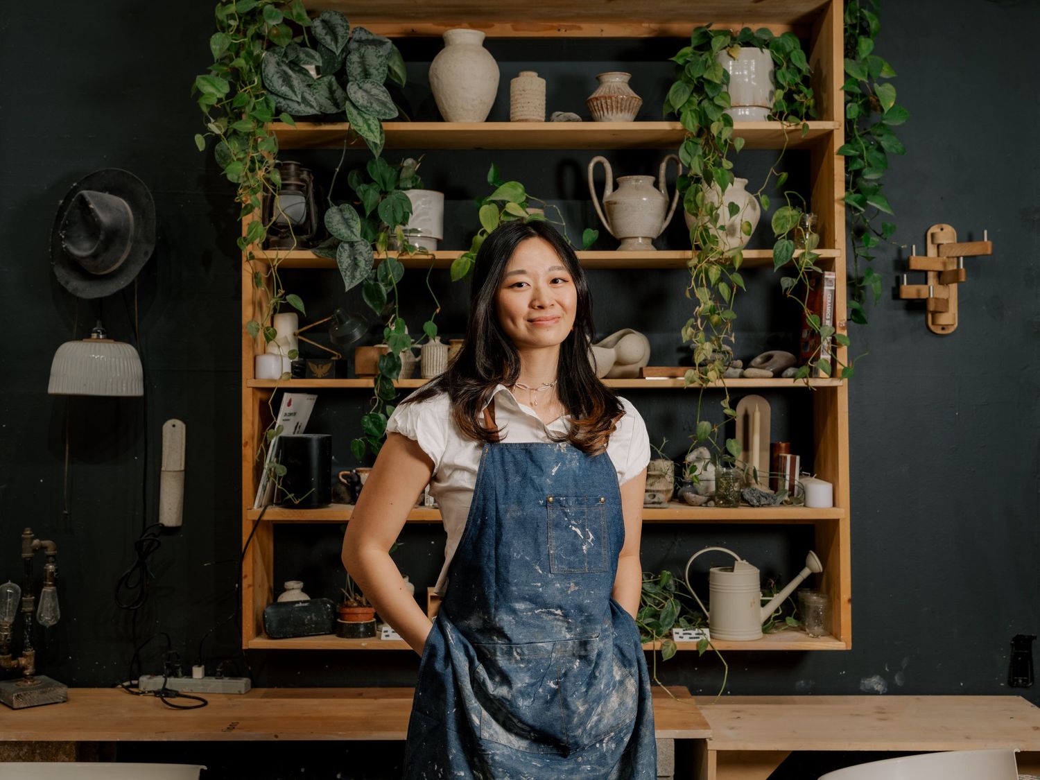 Ceramic pottery studio displays handmade vessels on wooden shelves with green plants and dark walls.