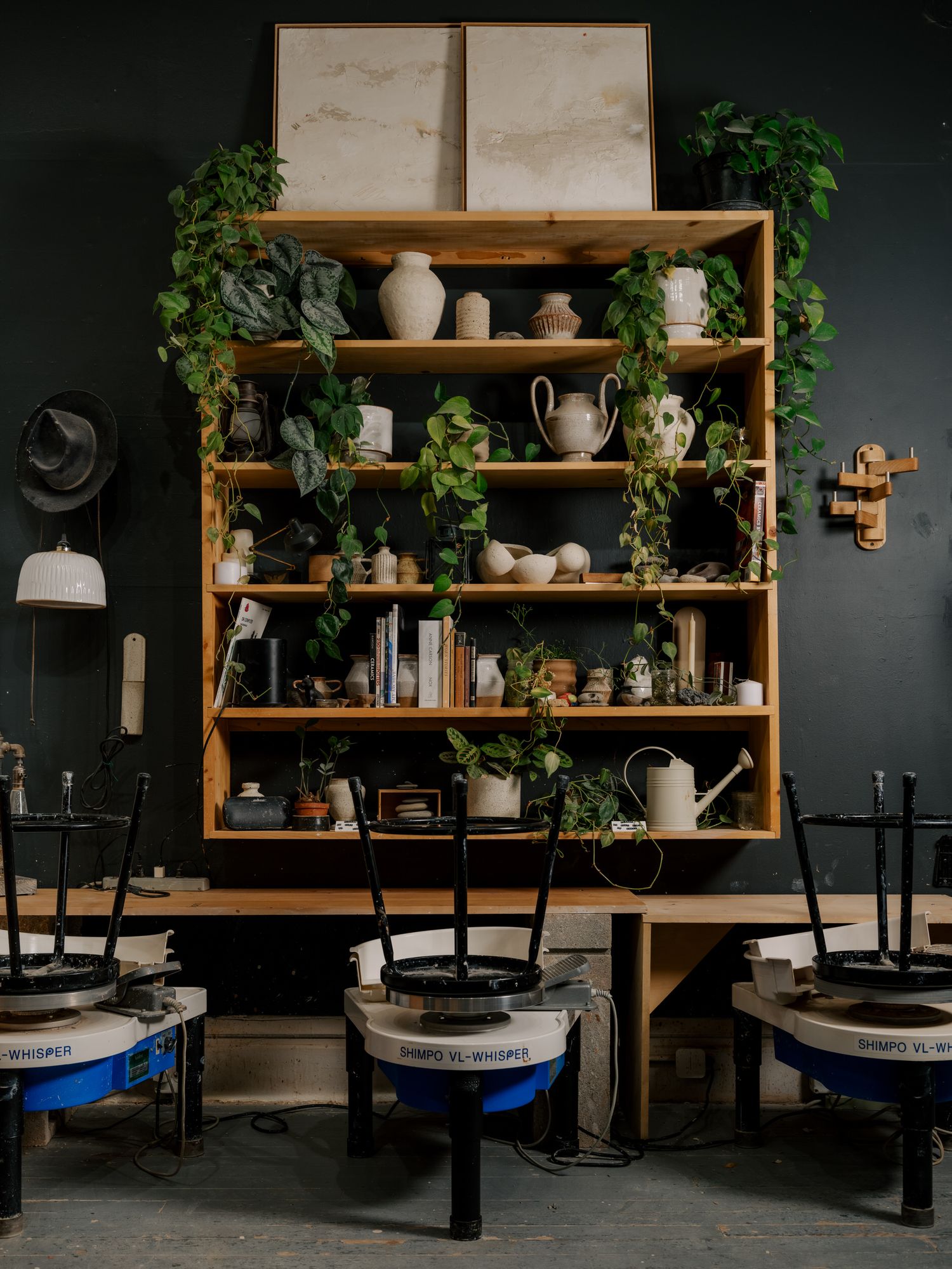 Pottery studio setup with wooden shelving unit displaying ceramics and trailing plants against dark wall above potter's wheels.