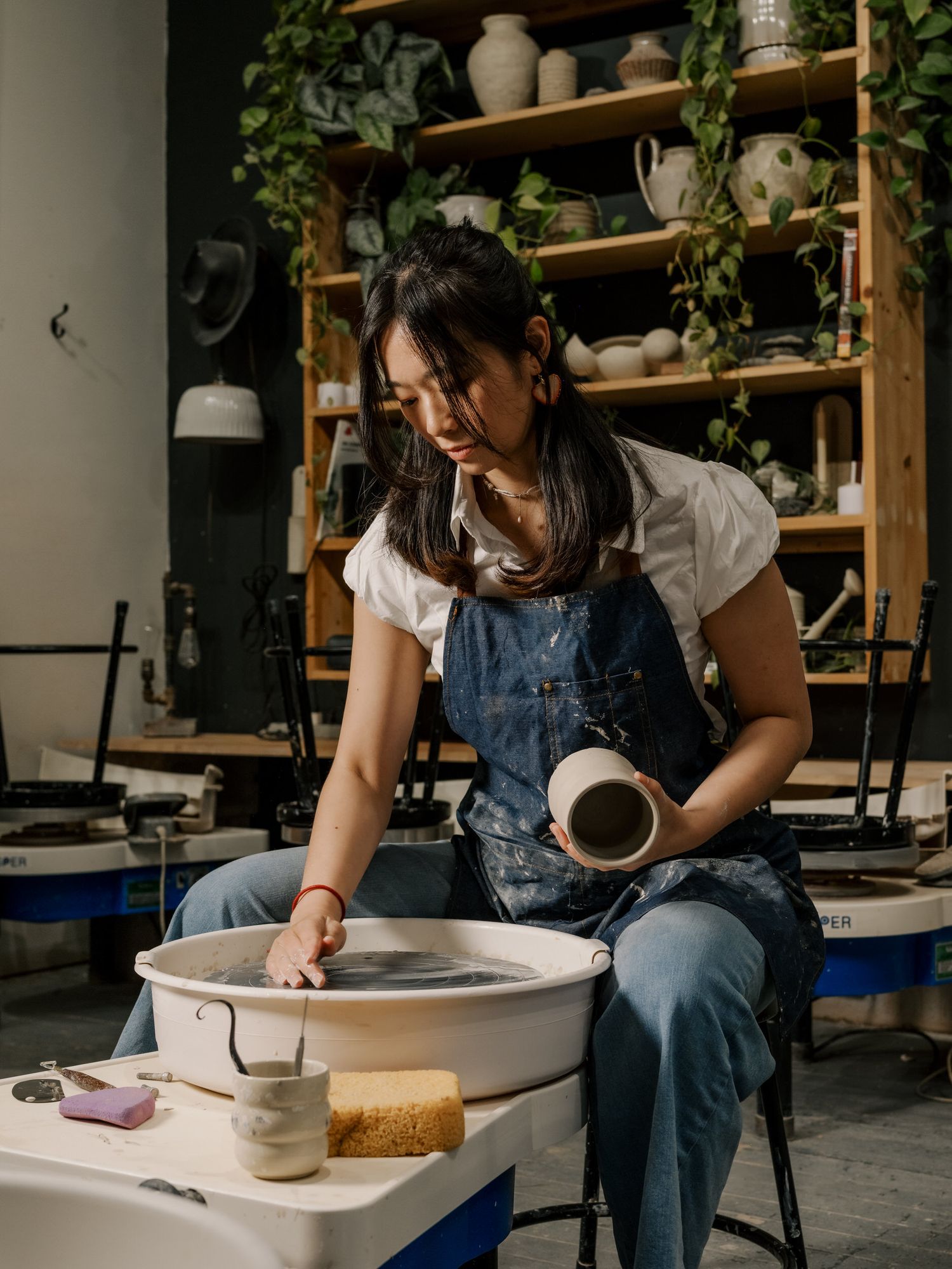 Artist in denim overalls works intently at a pottery wheel in a studio with wooden shelving.