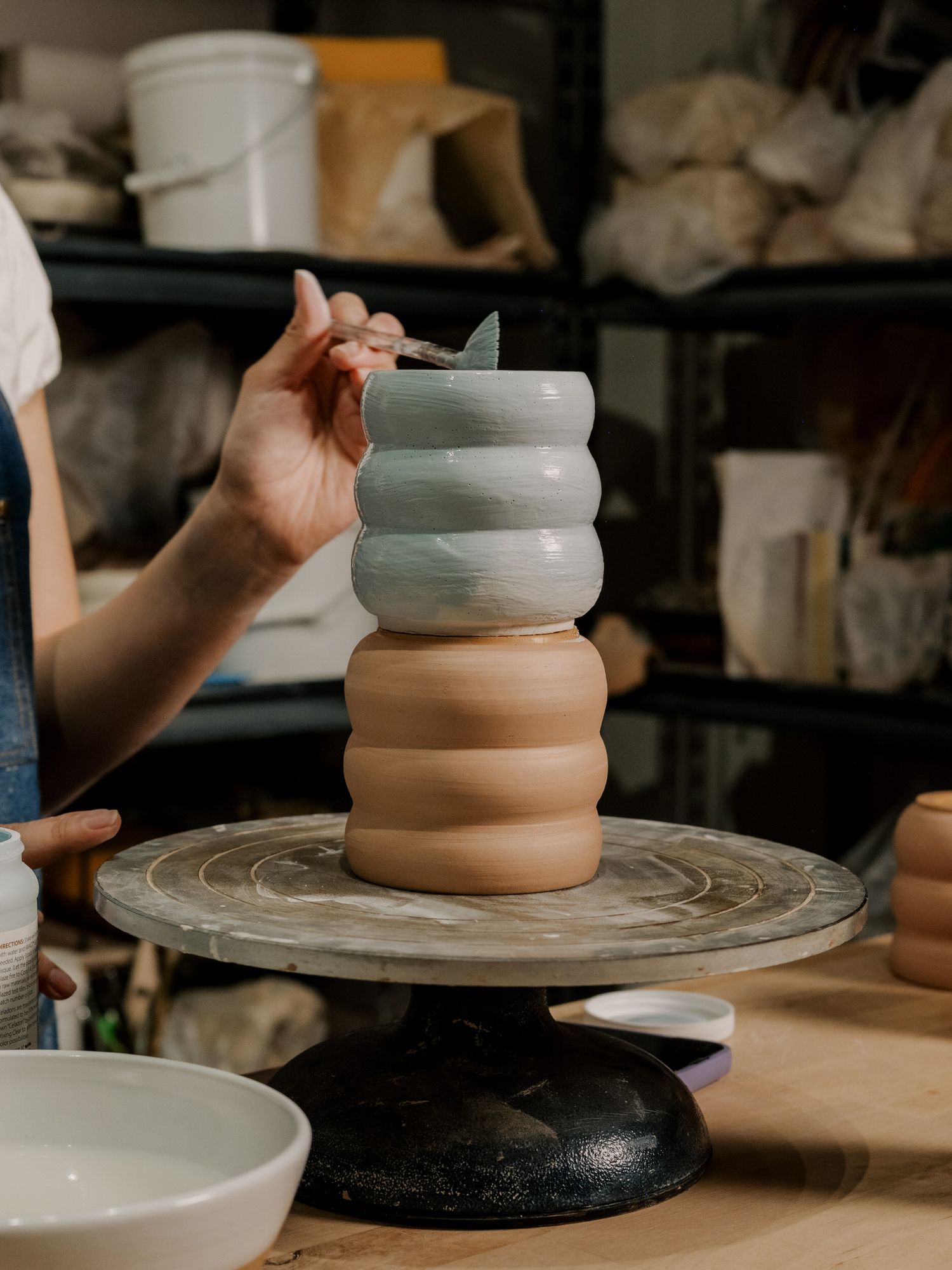 A hand shapes a ribbed ceramic vessel with alternating white and terracotta sections on a pottery wheel.
