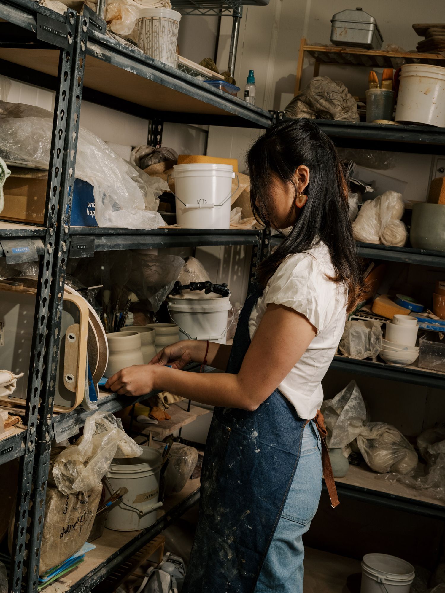 Artist working in pottery studio arranges ceramic pieces on industrial shelving units, showcasing creative workspace.