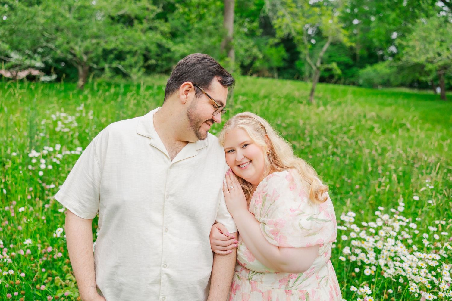 A couple sits together on a blanket in a sunny meadow filled with white wildflowers on a warm summer day.
