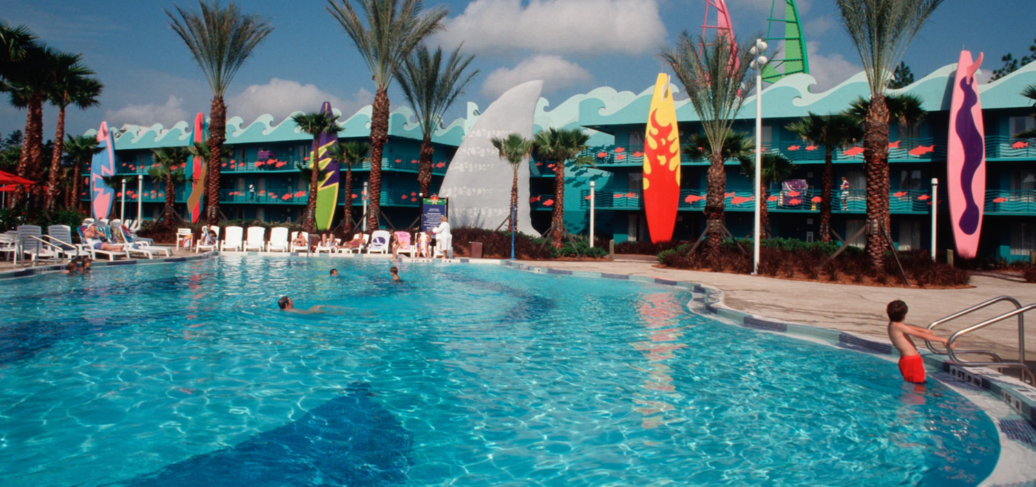 Resort swimming pool with surfboard decorations and palm trees against colorful hotel buildings.