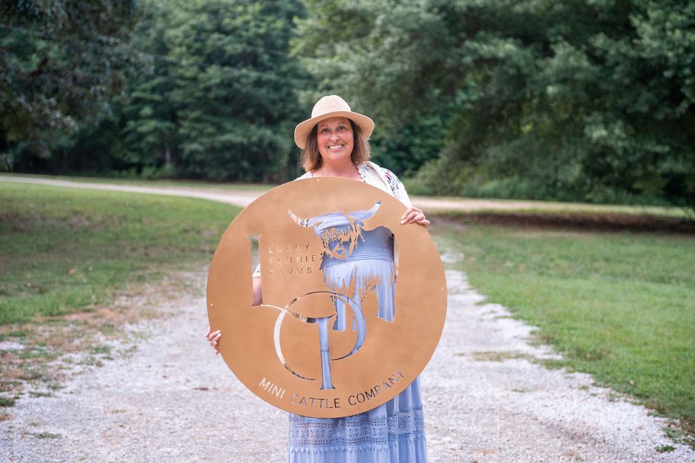 Person in blue gingham dress and sun hat holds large wooden circular sign on gravel path in park setting.