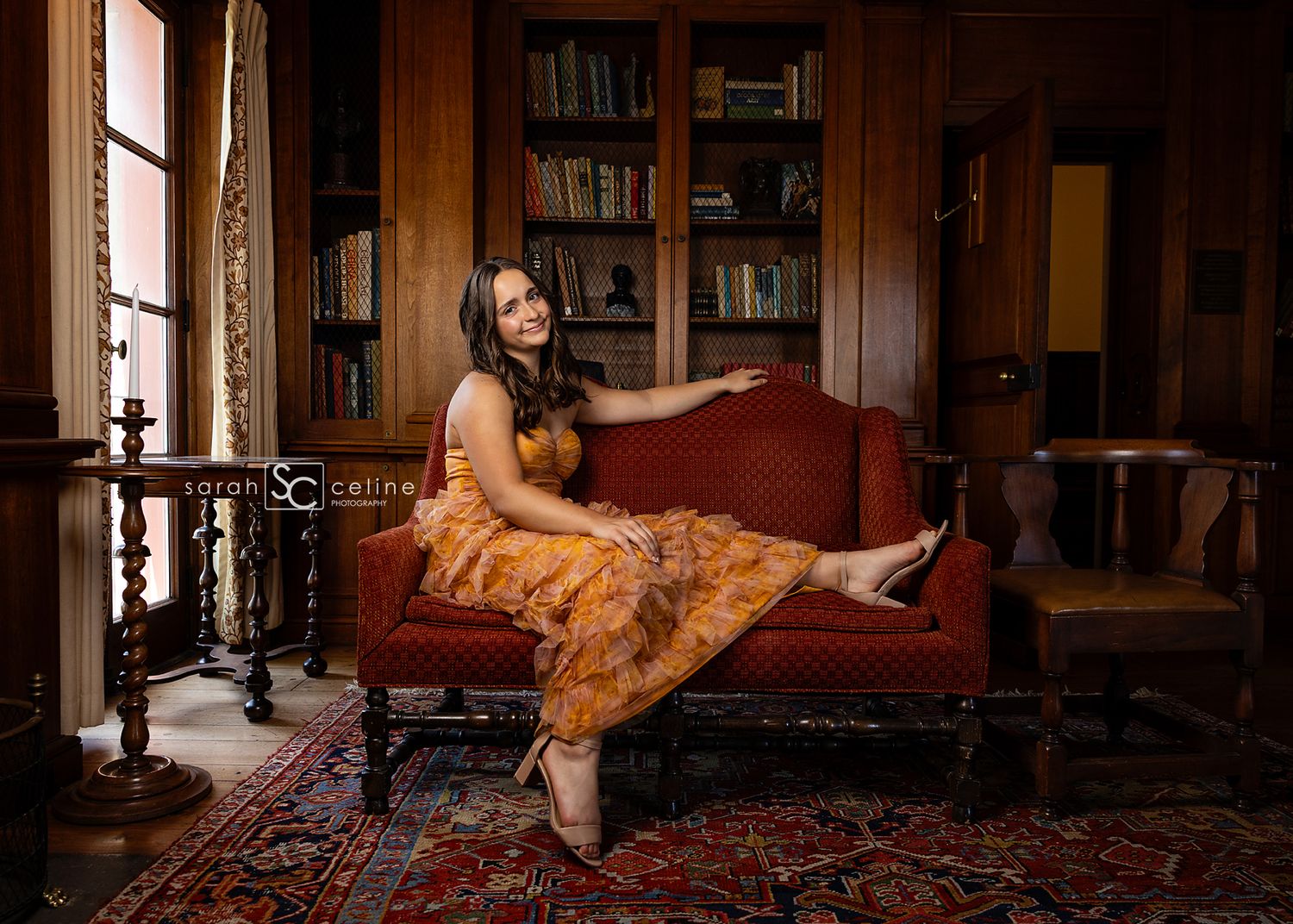 A graceful figure reclines on a vintage red couch in an elegant wood-paneled library with ornate decor and period details.