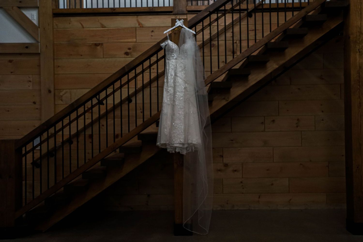 Wedding dress and veil hanging illuminated against wooden wall and staircase in rustic venue interior.