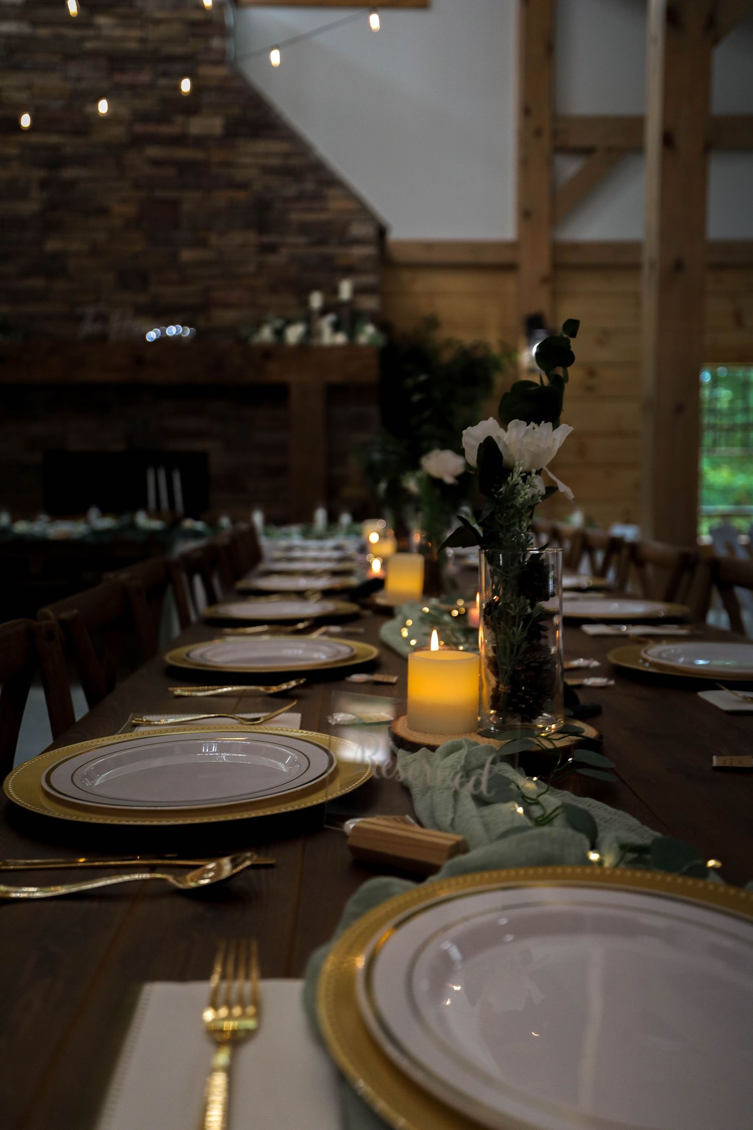 Elegant wedding reception table setting with gold charger plates, candlelight and floral centerpieces in rustic venue.