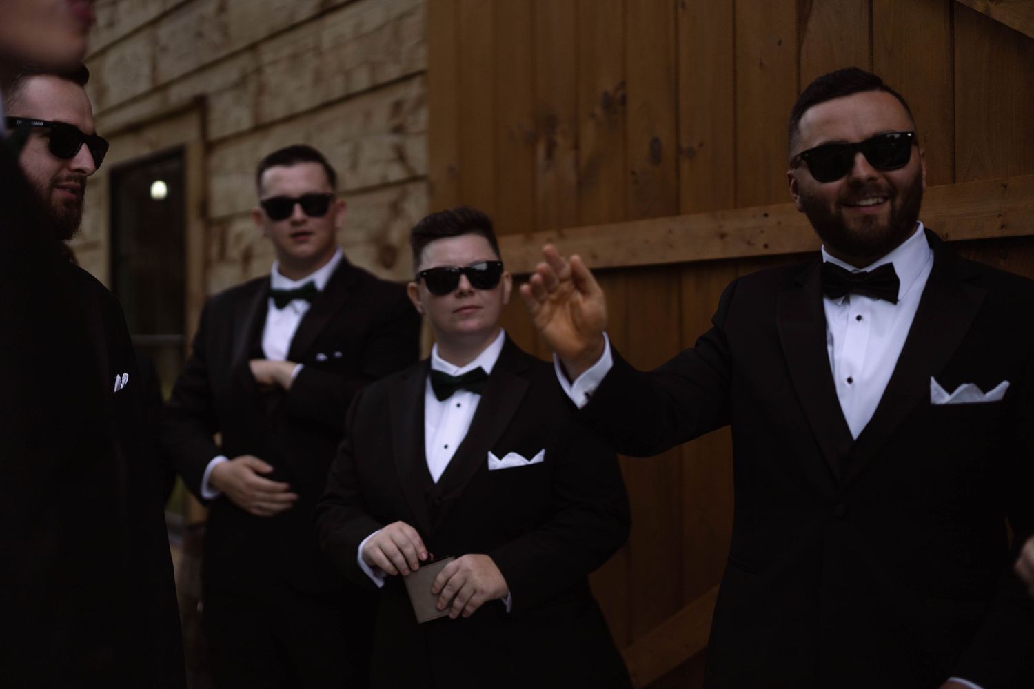 Several people wearing formal black tuxedos and sunglasses pose together in a traditional wedding party style.