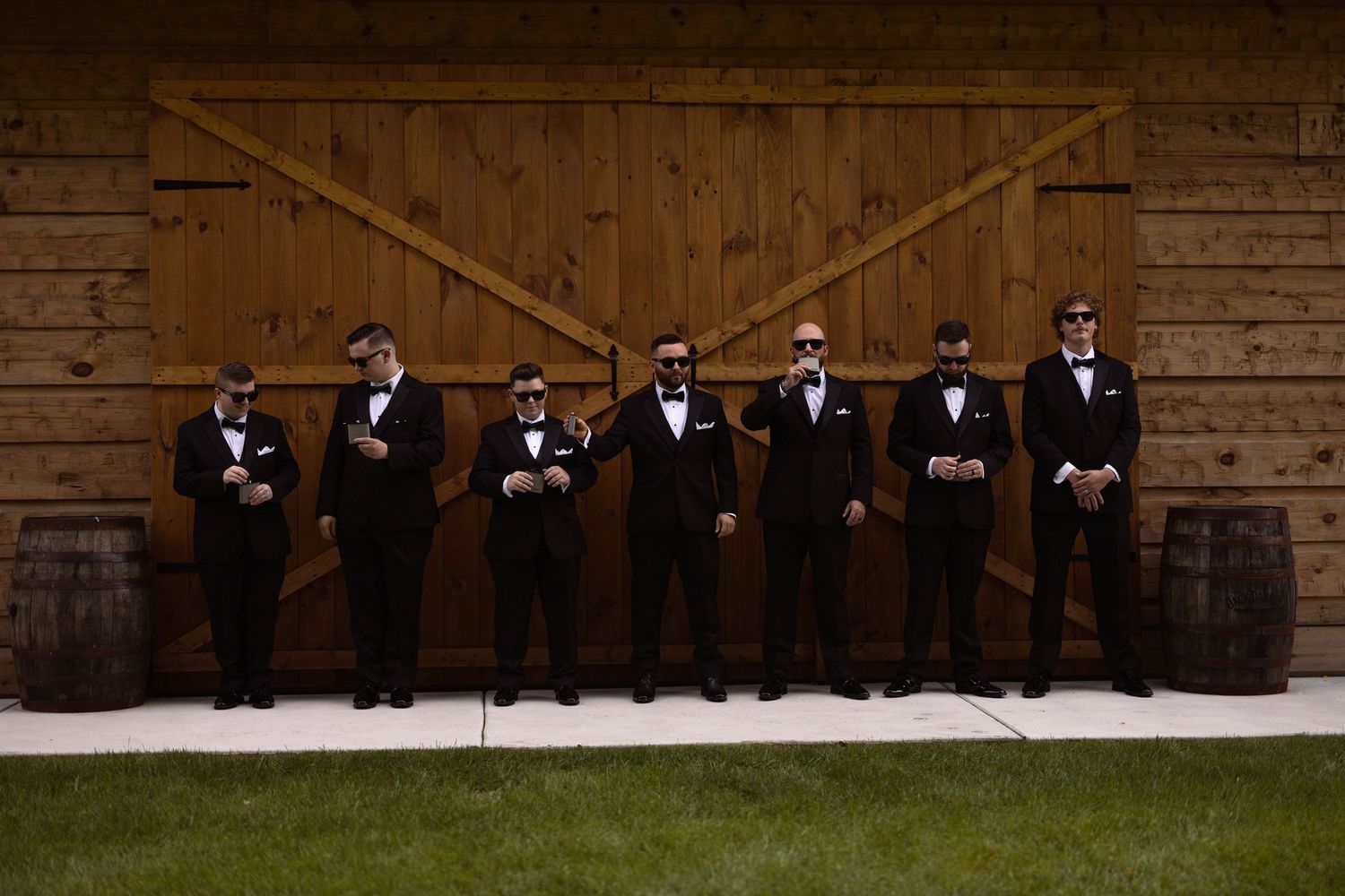 Groomsmen wearing black tuxedos sit casually on rustic wooden steps against a weathered barn wall.