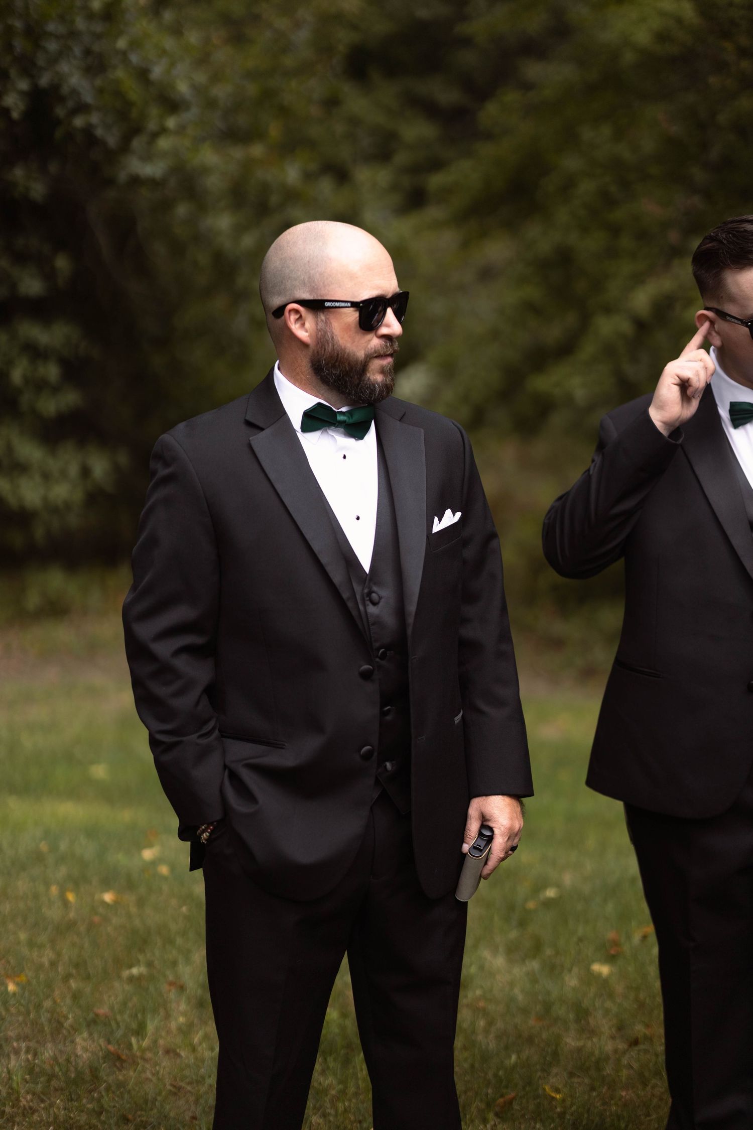 A groom in a black tuxedo and sunglasses stands outdoors during a wedding.