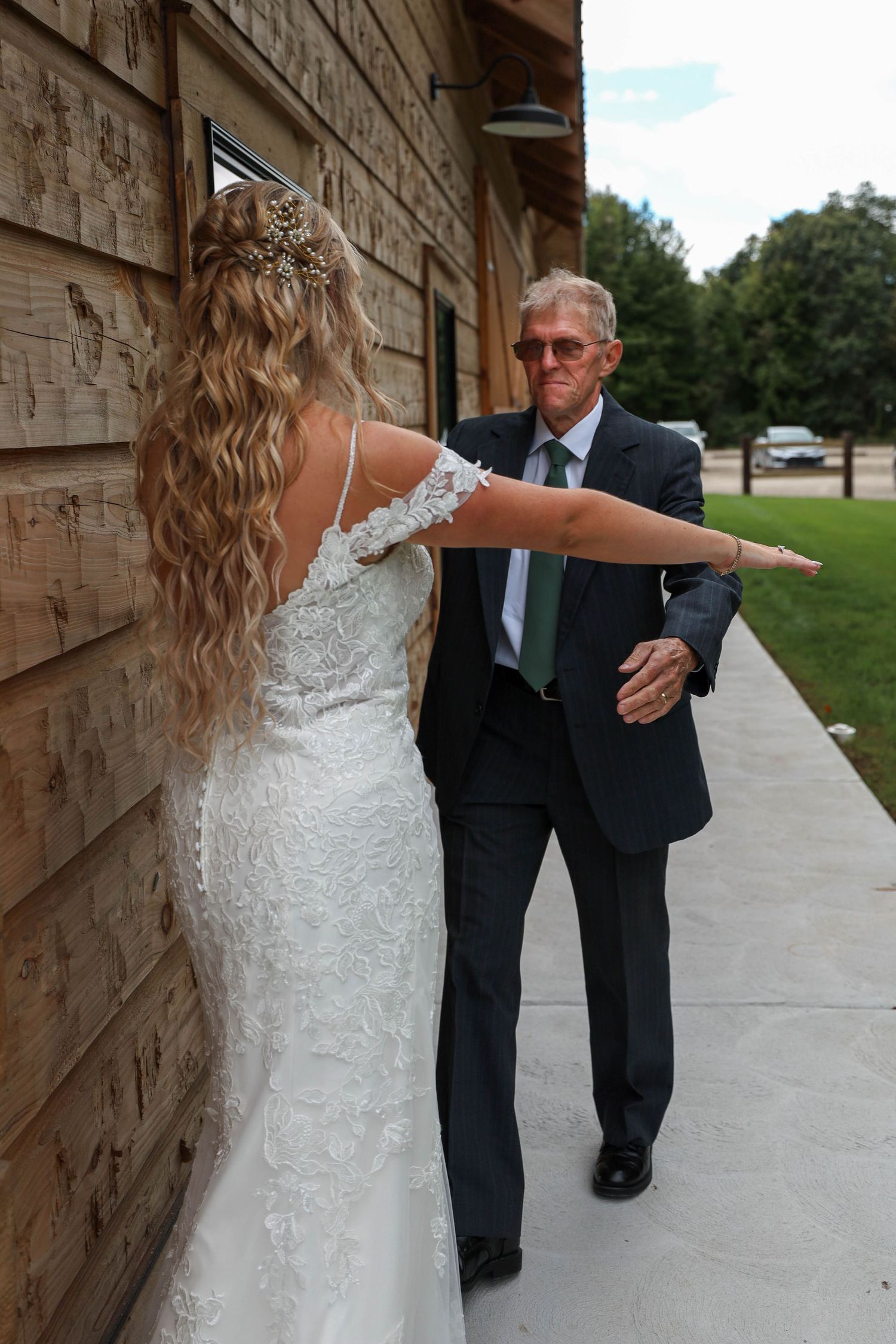 A bride in a white lace wedding dress stands next to a rustic wooden building wall while posing with someone in a suit.
