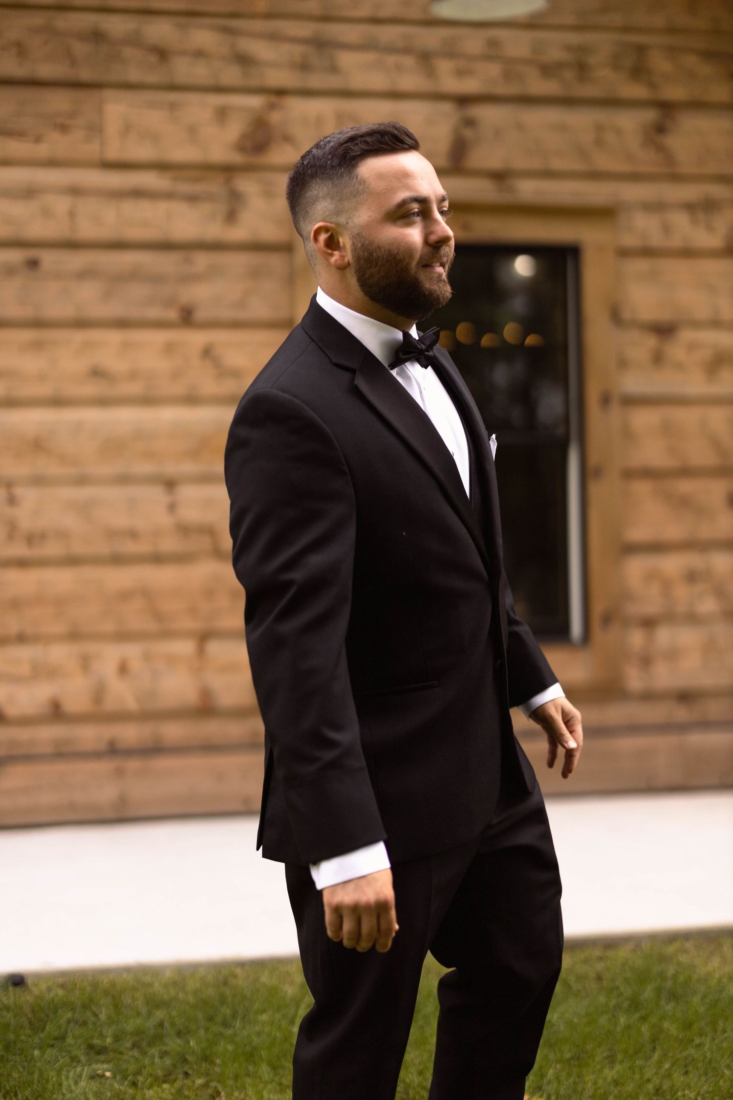 A groom in a classic black tuxedo smiles while standing against a rustic wooden wall.