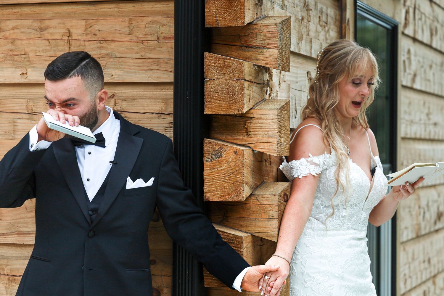 Bride and groom share an emotional first look while holding hands around a wooden corner wall.