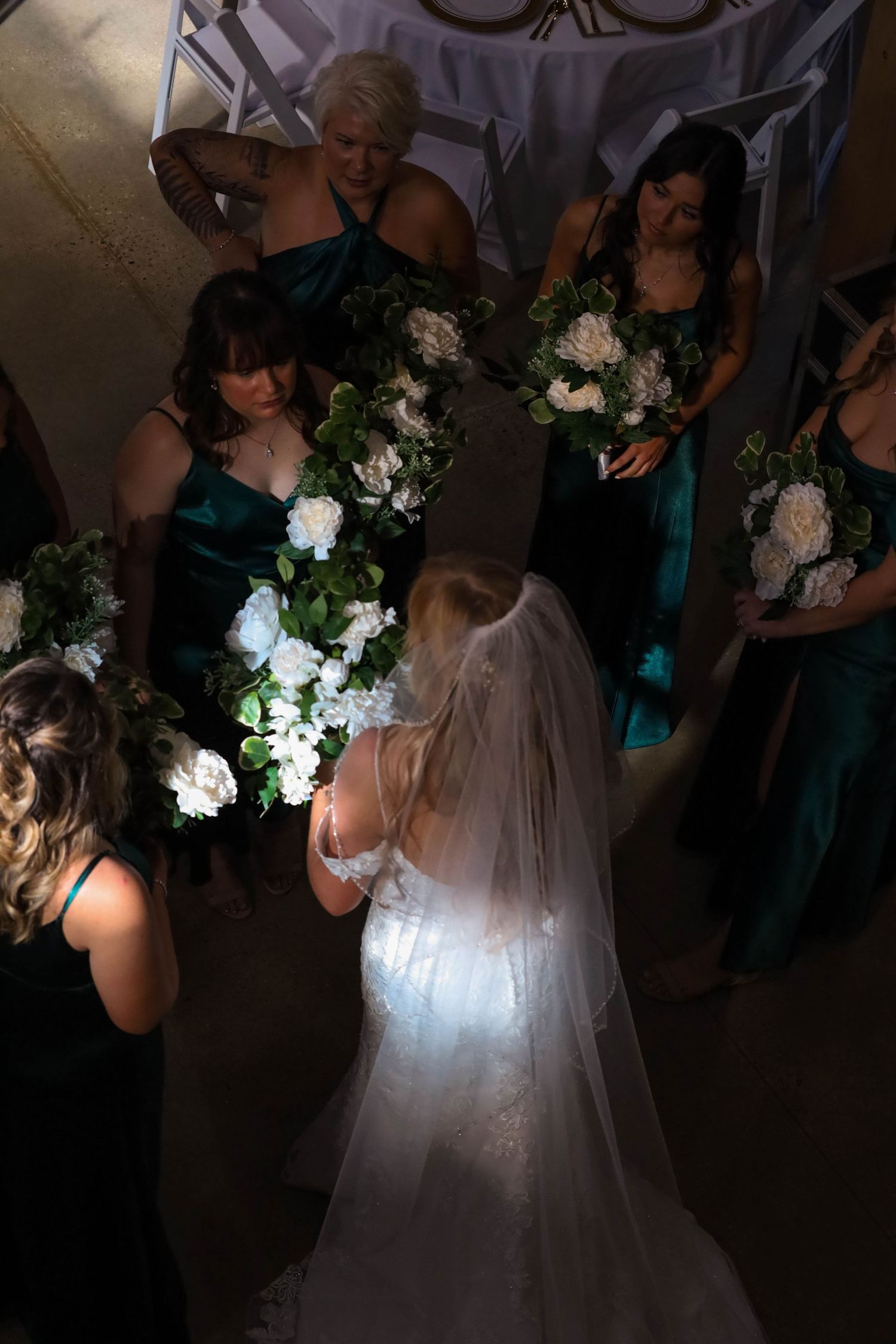 Bride in white wedding dress with veil surrounded by bridesmaids in teal dresses holding white floral bouquets.