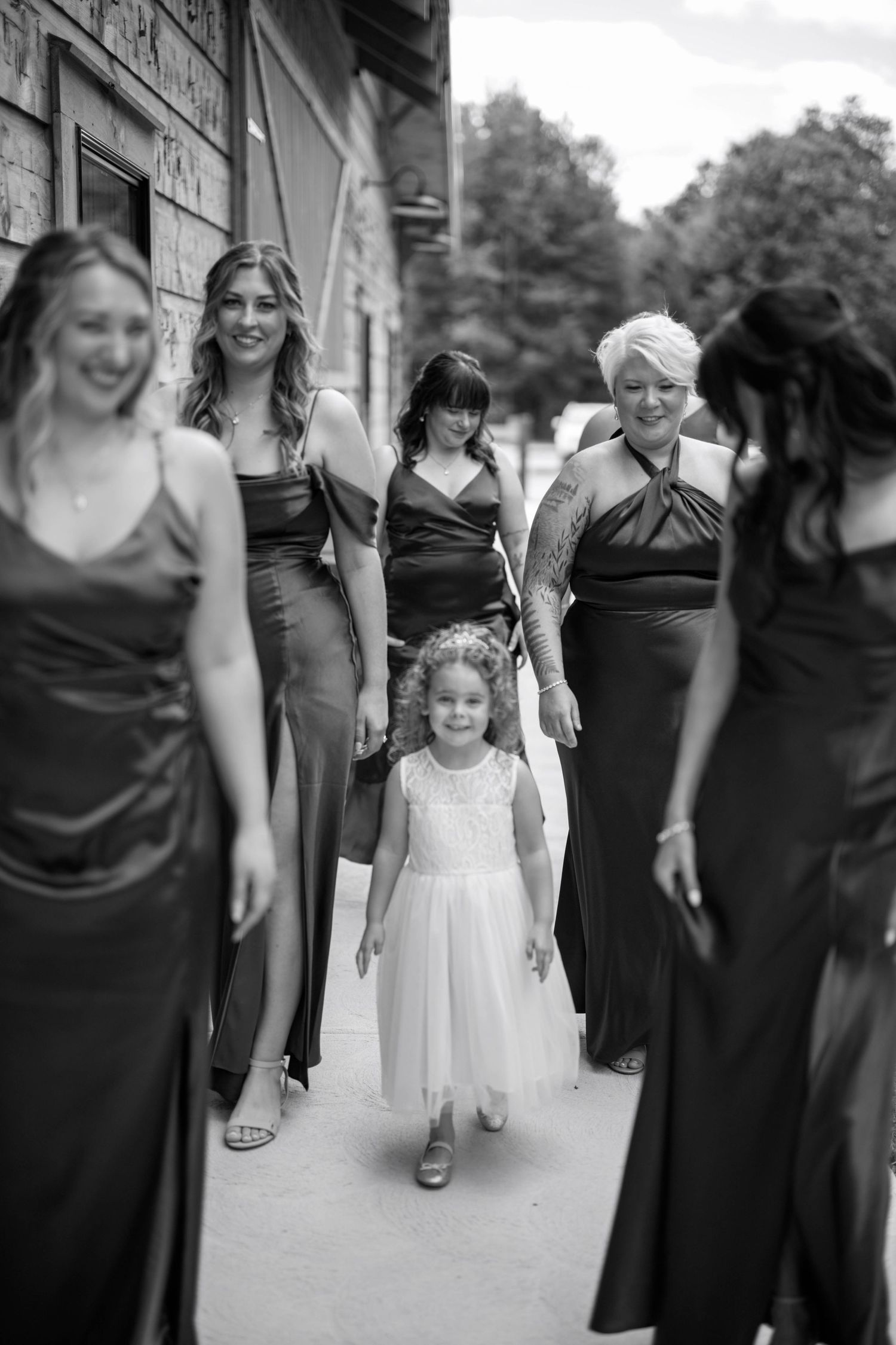 Young flower girl in white dress walking among bridesmaids wearing dark formal gowns.
