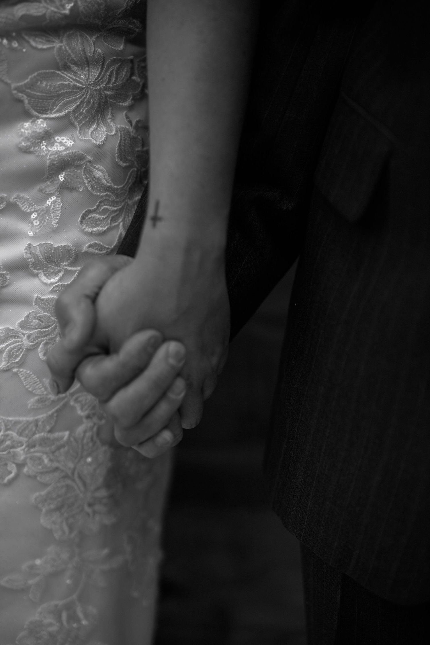 Black and white close-up of clasped hands with wedding dress lace details showing tender romantic moment.