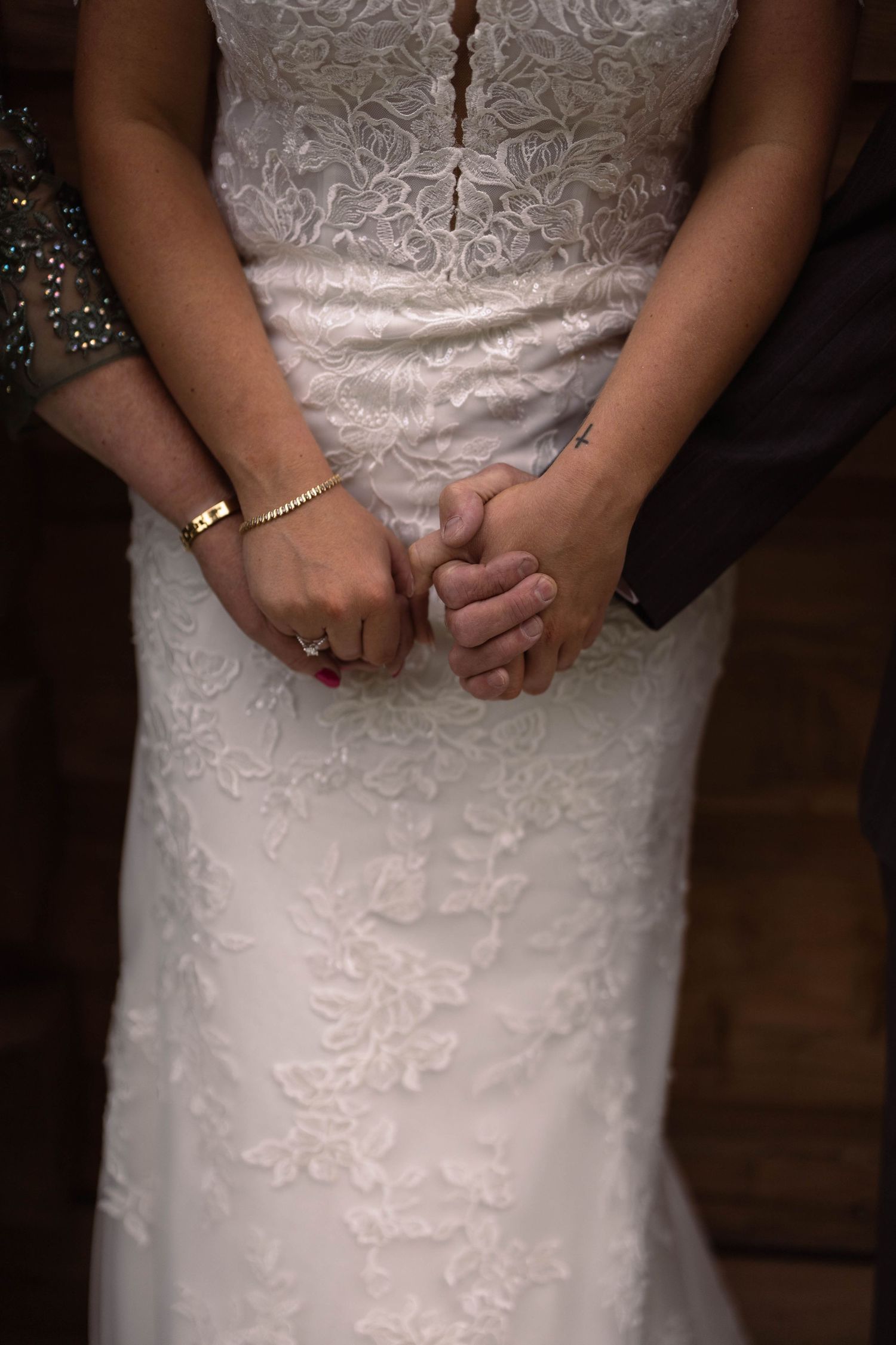 Close-up detail of hands clasped together wearing rings against a white lace wedding dress with floral embroidery.