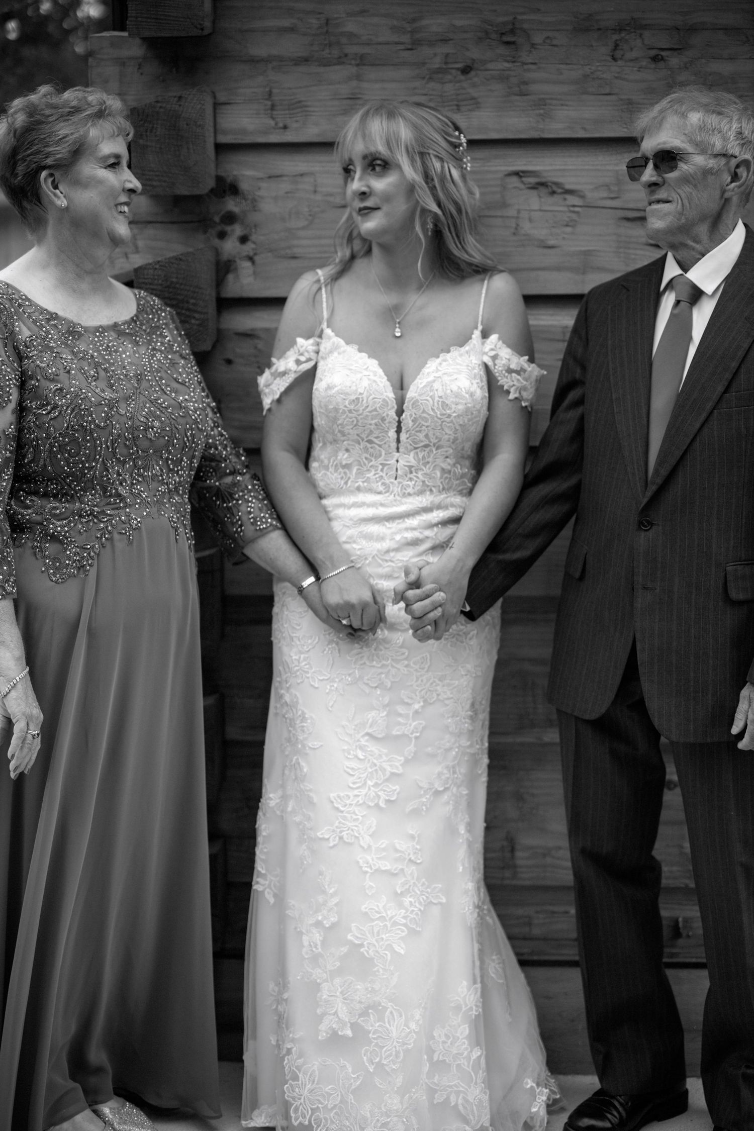 Black and white photo of a bride in a lace wedding dress standing with two guests in formal attire.