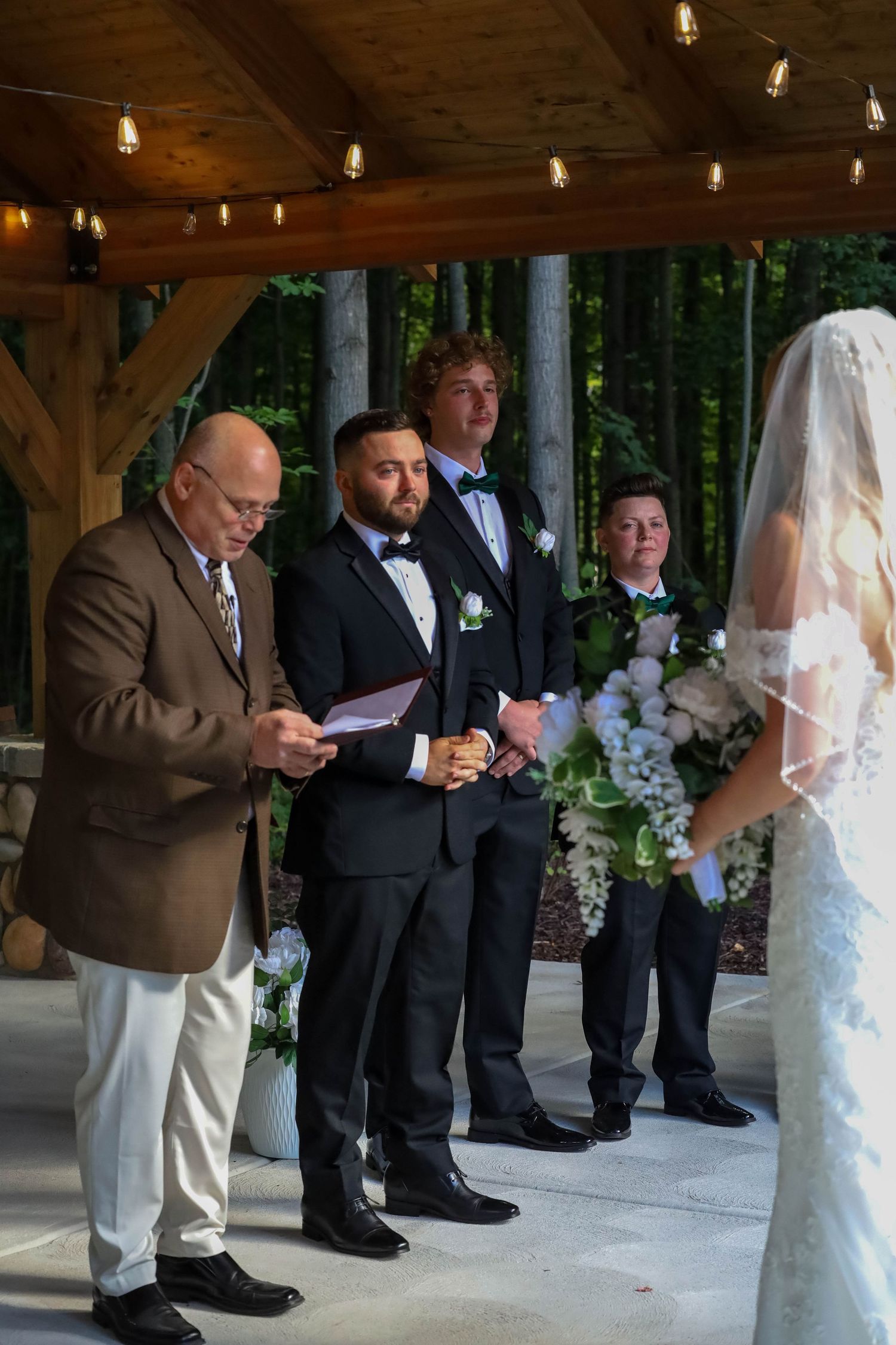 Wedding ceremony taking place under a covered outdoor pavilion with groomsmen and officiant.