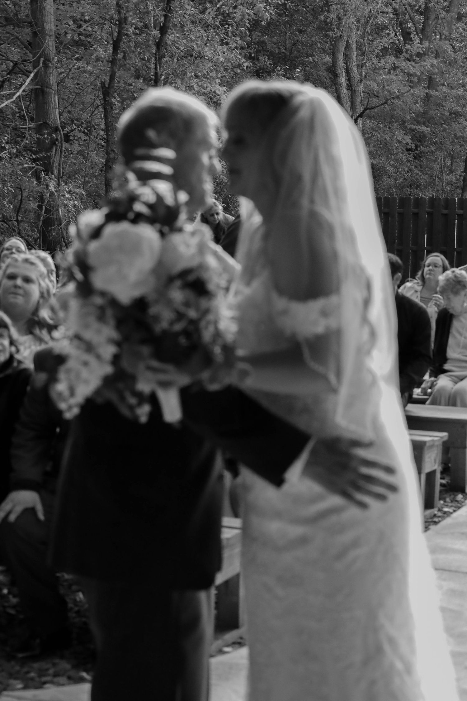 A romantic black and white outdoor wedding ceremony shows the bride in a veil holding a bouquet of flowers.