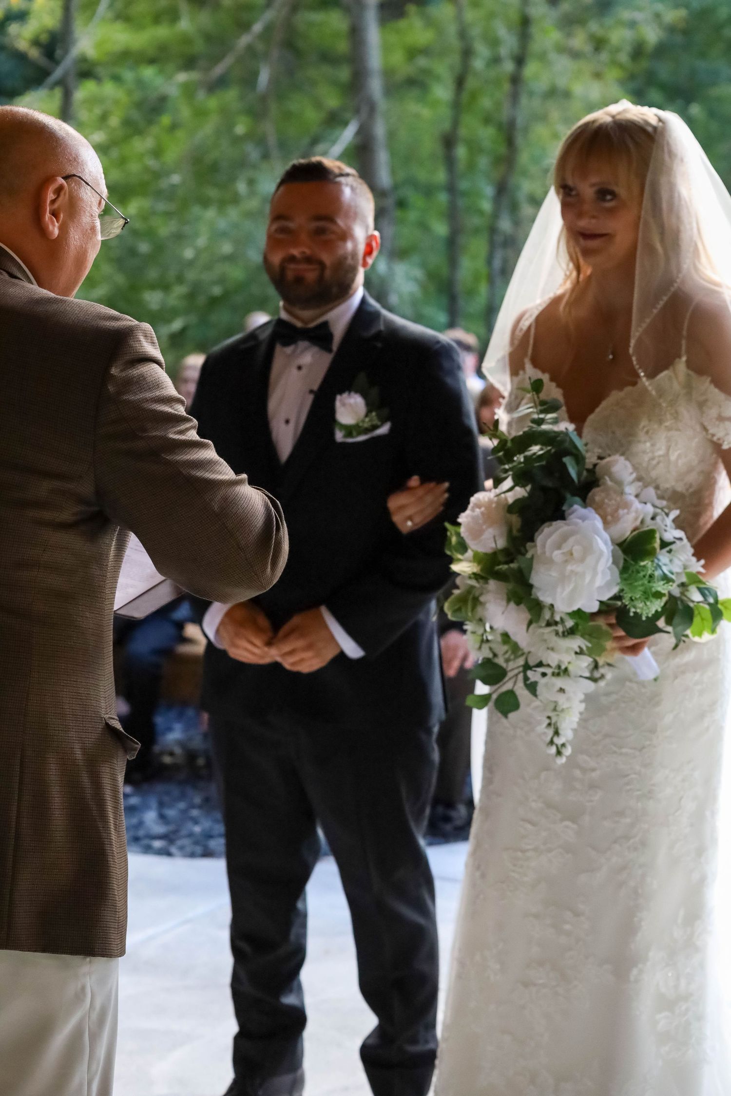 Wedding ceremony takes place outdoors with bride in white lace dress holding elegant white and pink floral bouquet.